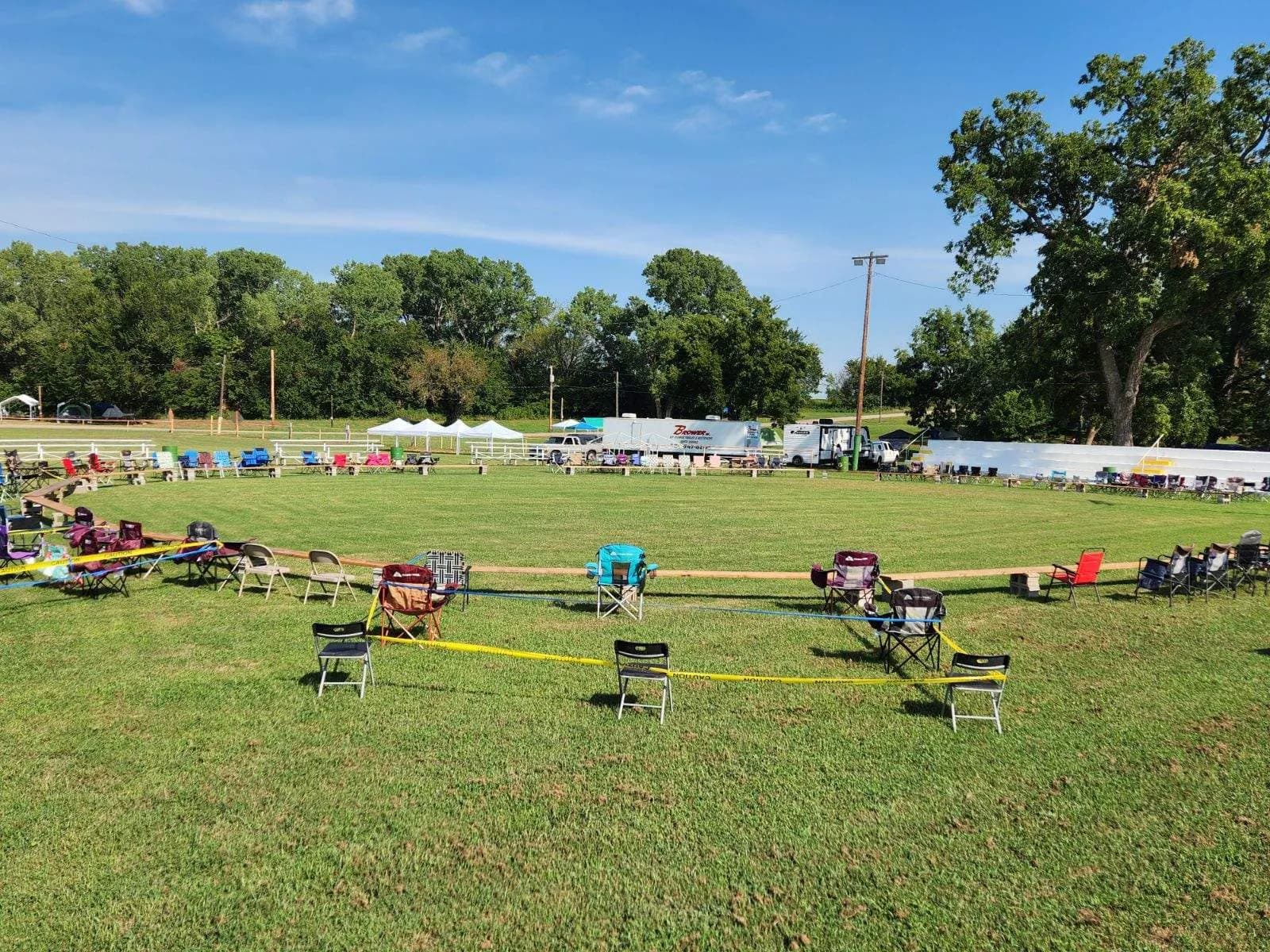 A Brower Inc. branded restroom trailer and white service truck parked along the far rail of a grassy Oklahoma community event arena, with lawn chairs set out around the ring and white vendor tents raised in the background before the crowd arrives